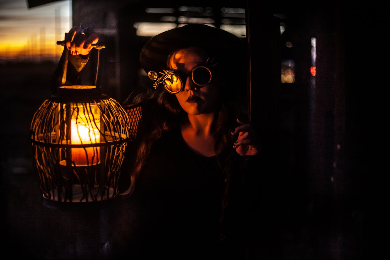 A black and orange composition showing a girl with steampunk goggles holding a caged lantern. The orange lantern is the only light source as the image quickly fades to black.