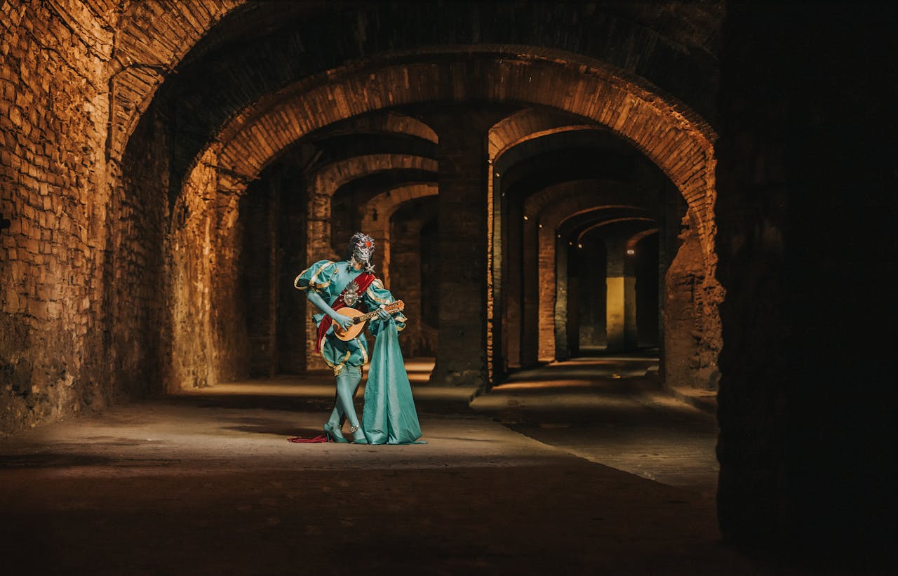 The setting is an arched stone walkway where stones of golden-brown are crossed by deep shadows. In the center, an ornately-dressed bard in turquoise strikes a dramatic pose with their lute or mandolin.