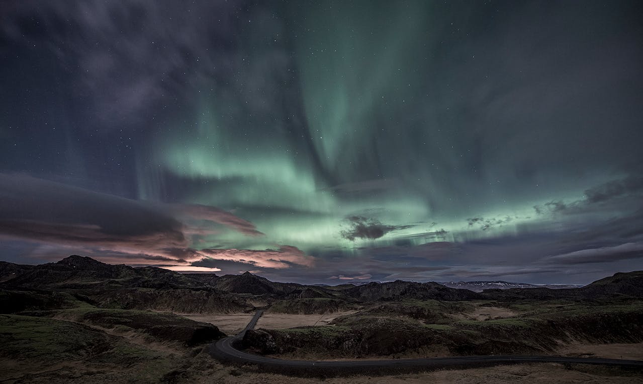 A green aurora over brown mountains with a bit of pink sunset on the clouds between