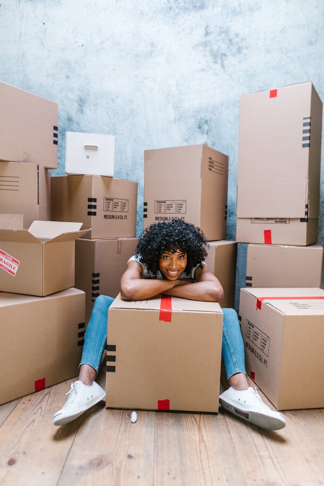 A black woman with curly hair and jeans rests her arms on top of a moving box taped with red tape, smiling at the camera. Behind her, boxes of various sizes are stacked.