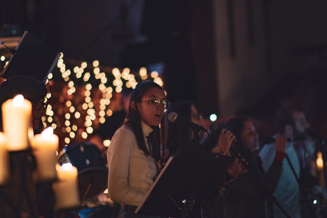 Women singing a song in a line in a church or somewhere similar. Only the first woman is in focus. There are dozens of lit candles behind and around them.