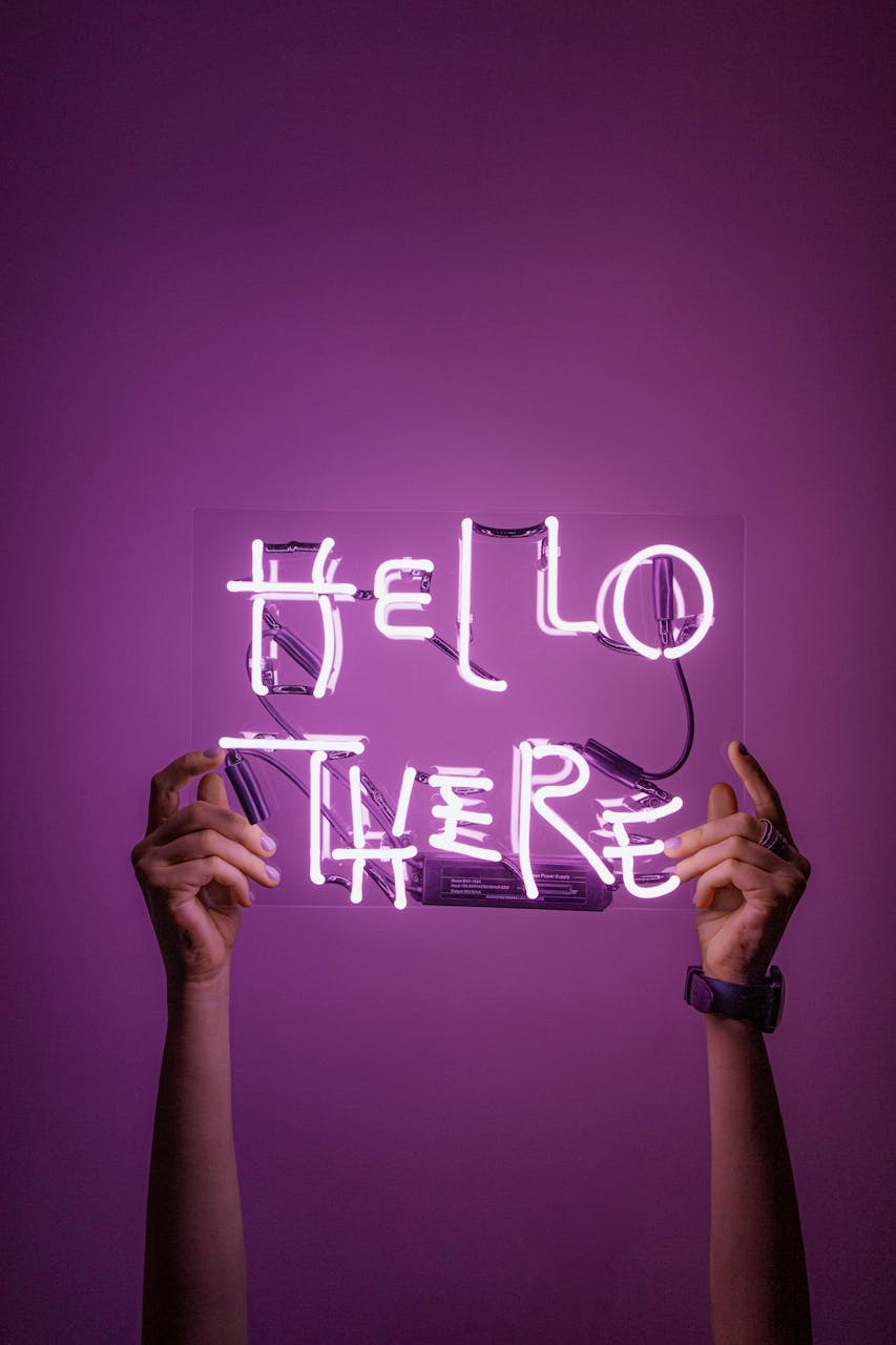 Against a purple background, two hands hold up a purple neon sign that says Hello There