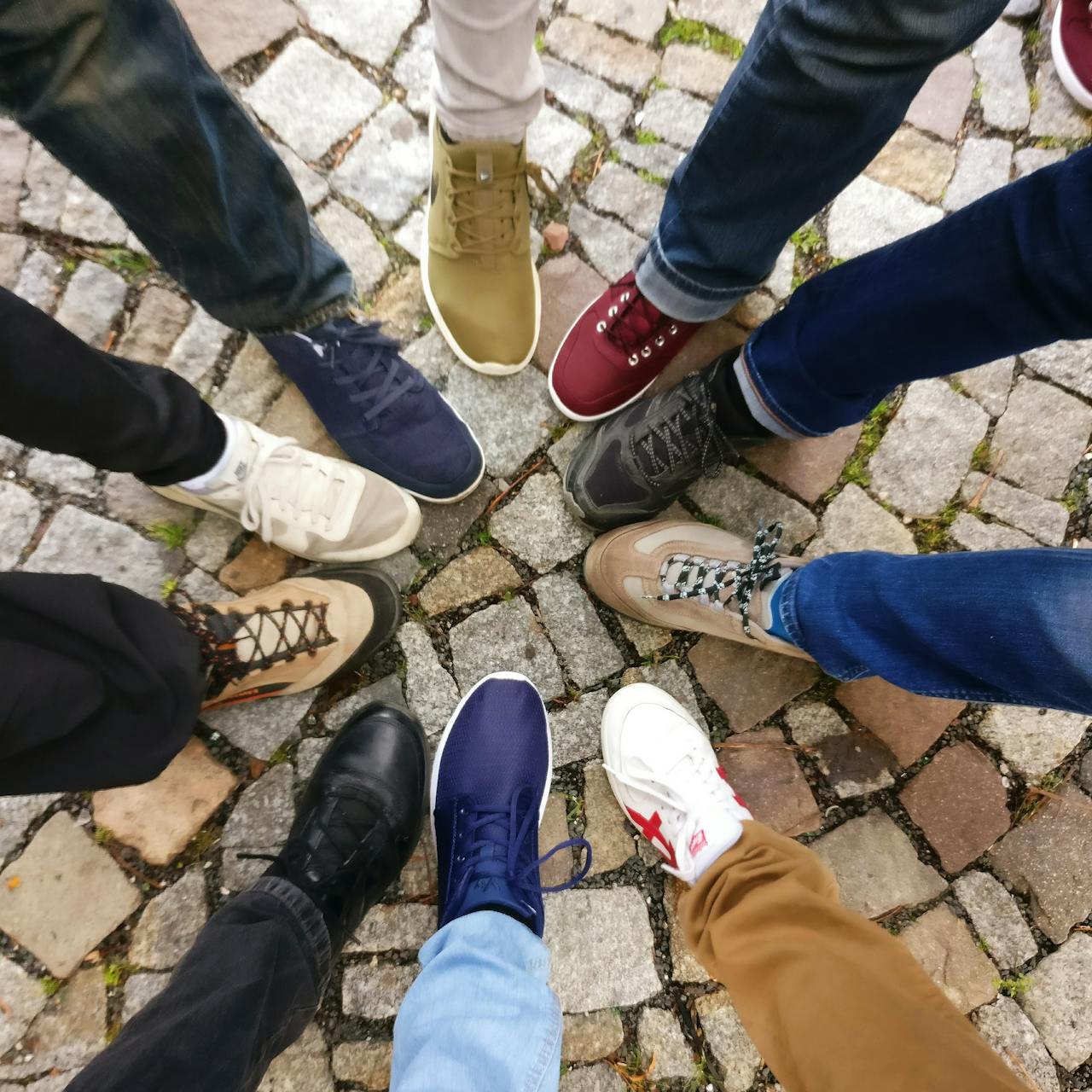 A circle of people standing on a stone walk have each put one foot in to the middle of the circle. The shoes are varied in color and style.